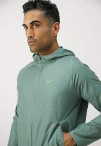 Veste légère à capuche vert clair avec fermeture éclair sur le devant. Comprend un petit logo Nike gris sur la poitrine et des poches latérales. Texture lisse.