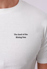 T-shirt blanc avec un petit texte noir « La Terre du Soleil Levant » centré sur le haut de la poitrine, porté par une personne à la peau de ton moyen.