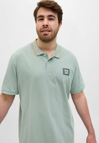 Heled roheline polo särk, mis on valmistatud puuvillast, kolmesuunalise nööbiplahvi, oranžide kaeluse aktsentide ja väikese logoplaadi ning rinnakilikla. 