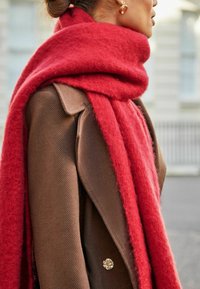 Rød uldscarf draperet omkring en brun tekstureret frakk, der har en dekorativ knap. Scarfet har en blød, fuzzy tekstur og en bred silhuet.