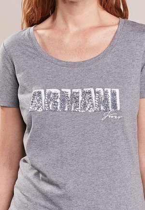 Kobieta nosząca szary t-shirt z dużymi, srebrnymi cekinowymi literami tworzącymi napis „ARMANI” oraz mniejszym białym napisem „Jeans” poniżej.