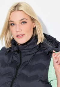 Schwarze Steppweste mit hochgeschlossenem Kragen und Reißverschluss, mit einer strukturierten Oberfläche. Das Model trägt darunter ein hellgrünes Hemd.