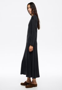 Vestido largo de terciopelo gris oscuro con mangas largas y un corte relajado, que presenta una ligera forma en A y un escote redondo. Llevado con zuecos marrones.