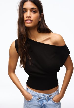 Mladá žena s dlhými hnedými vlasmi oblečená v čiernom jednoramennom topu a svetlomodrých džínsoch, ruky v kapsách, na bielom pozadí.