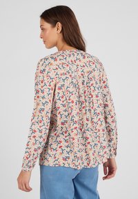 Blusa floreale con sfondo beige, caratterizzata da motivi rossi e blu, maniche lunghe e un dettaglio arricciato sul retro per una maggiore texture.
