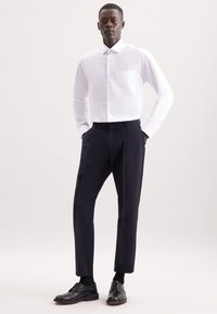 Man staat met zijn handen in zijn zakken, gekleed in een witte knop-up shirt, zwarte op maat gemaakte broek, zwarte sokken en zwarte schoenen op een effen achtergrond.