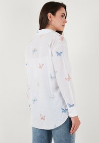 White long-sleeve shirt featuring a butterfly pattern in blue and pink, with a collar and buttoned cuffs. Made from lightweight fabric.