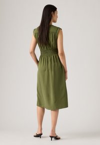 Olivza zaļa kleita ar pieguļošu, smockētu jostasvietu. Īsas piedurknes, ceļgala garuma siluets un mīksta tekstūra. Valkāta ar melniem augstpapēžu sandaliem.