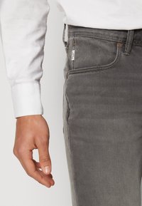Höger hand av en person som bär en vit långärmad skjorta och grå denimjeans med synliga sidofickor och sömnadsdetaljer.