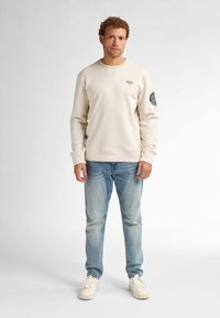 Felpa beige con scollatura rotonda, polsini a costine e grafiche sul petto; abbinata a jeans azzurri chiari e sneakers bianche.