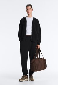 Tânăr purtând o hanorac negru cu fermoar, tricou alb, pantaloni negri și adidași, ținând o geantă de voiaj maro mare de o curea.