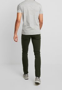 Mężczyzna odchodzący, ubrany w szary krótki rękaw t-shirt, ciemnozielone spodnie o dopasowanym kroju i szare trampki na neutralnym tle.