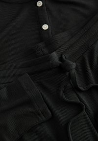 Schwarzer gerippter Stoff mit strukturierten Oberflächen. Verfügt über eine Knopfleiste mit weißen Knöpfen und eine gebundene Kordel am Bund.