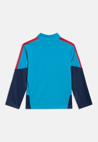 Top atlético de mangas compridas em azul vibrante com detalhes vermelhos nos ombros, mangas azuis com padrão e gola alta; textura suave.