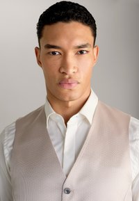 Helles rosa Weste mit feiner Textur, über einem weißen Hemd getragen. Verfügt über einen Knopfverschluss und eine maßgeschneiderte Passform.