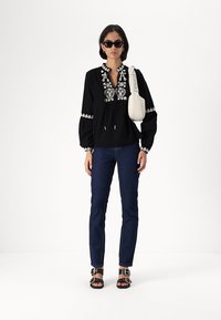 Μαυροσ μπλούζα με μακριά μανίκια και άσπρη λουλουδάτη κεντητική, συνδυασμένη με μπλε εφαρμοστά τζιν, μαύρα σανδάλια με χρυσές λεπτομέρειες και άσπρη τσάντα.