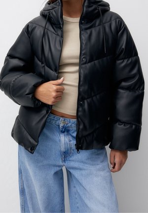 Personne portant une veste noire matelassée à capuche sur un top court côtelé beige et un jean large bleu clair, avec une main ajustant la veste.