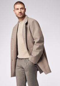 Hellbrauner Wollmantel mit Reverskragen, getragen über einem beigen, gerippten Pullover und maßgeschneiderten grauen Hosen mit lockerem Schnitt.