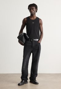 Top fără mâneci negru cu logo-ul "HUGO", blugi negri din denim, curea neagră și mocasini negri. Modelul ține o geantă din piele neagră. Design simplu.