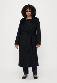 Cappotto lungo nero con design avvolgente, cintura a nastro, due tasche frontali, rever ampi e tessuto liscio. Indossato sopra un top e pantaloni neri.