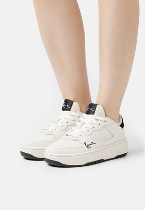 Pieds portant des baskets basses blanches avec des accents noirs au talon et à la languette, des bords de semelle noirs, et un détail emblématique sur les côtés et la languette.