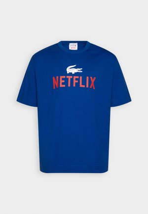 T-shirt bleu à manches courtes avec le logo crocodile blanc de Lacoste au-dessus du mot « NETFLIX » en lettres capitales rouges sur la poitrine.