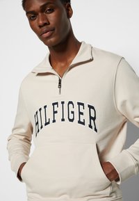 Felpa cremosa con zip e logo "HILFIGER" in blu scuro in evidenza. Materiale morbido con tasca a marsupio e polsini a coste.