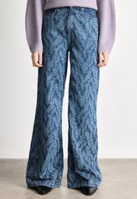 Άτομο που φοράει ψηλόμεσα φαρδιά τζιν με μπλε και λευκό αφηρημένο σχέδιο, πλεκτό πουλόβερ λεβαντίνι και μαύρα μυτερά παπούτσια.