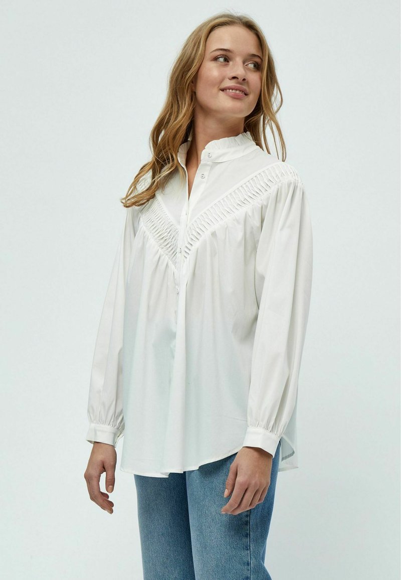 Camicia bianca con bottoni, caratterizzata da un design plissettato sul giogo, maniche lunghe e colletto alto, realizzata in tessuto liscio.