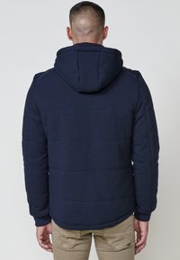 Giacca a vento blu navy con superficie texture, cappuccio e polsini a costine. Presenta cuciture orizzontali e una silhouette aderente.
