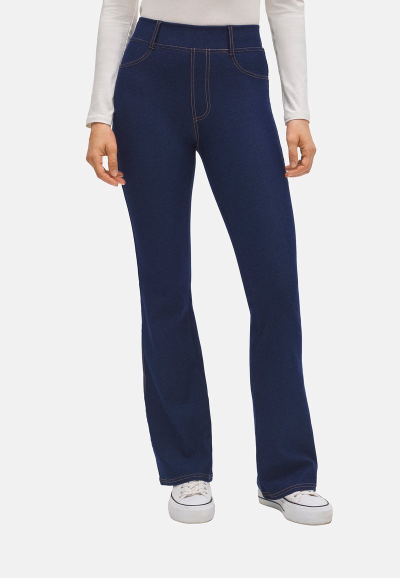 Vaqueros de campana azul marino con cintura alta, que cuentan con bolsillos frontales y costuras en contraste de color naranja. Textura de denim suave. Llevados con zapatillas blancas.