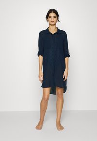 Navy button-up dress with two front pockets and roll-up sleeves. Made of lightweight fabric with a textured diamond pattern.