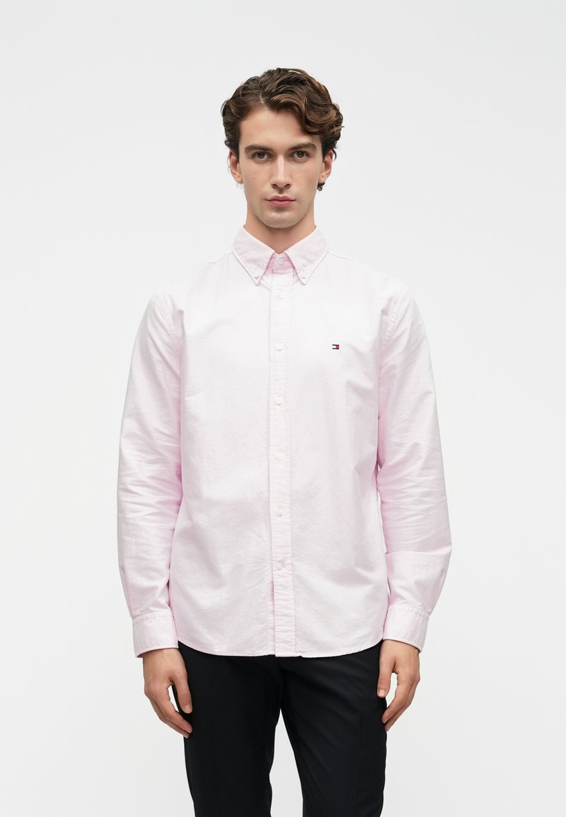 Gaiša rozā pogājamā krekla ar klasisko apkakli, garām piedurknēm un mazu logotipa detaļu. Audums izskatās gluds un viegls.