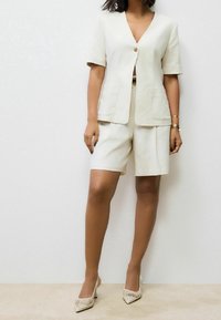 Licht beige pak met een blazer met korte mouwen, een V-hals, een enkele knoopsluiting en twee voorzakken; gecombineerd met bijpassende shorts en schoenen.