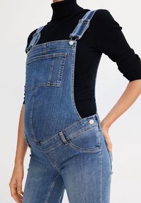 Denim overalls in light blue with adjustable straps, front pocket, and smooth texture, designed for a fitted style over a black top.