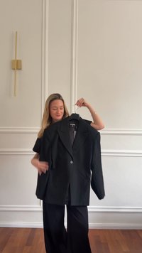 Musta bleiseri, jossa on käännetty kaulus, yksittäinen napitus, kaksi etutaskua ja löysä istuvuus, esillä henkariin ripustettuna yksinkertaista seinää vasten.