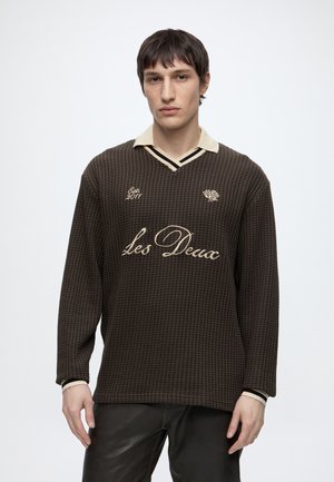 Giovane uomo che indossa un maglione marrone lavorato con collo beige e scritta "Les Deux", abbinato a pantaloni neri, in piedi davanti a uno sfondo semplice.