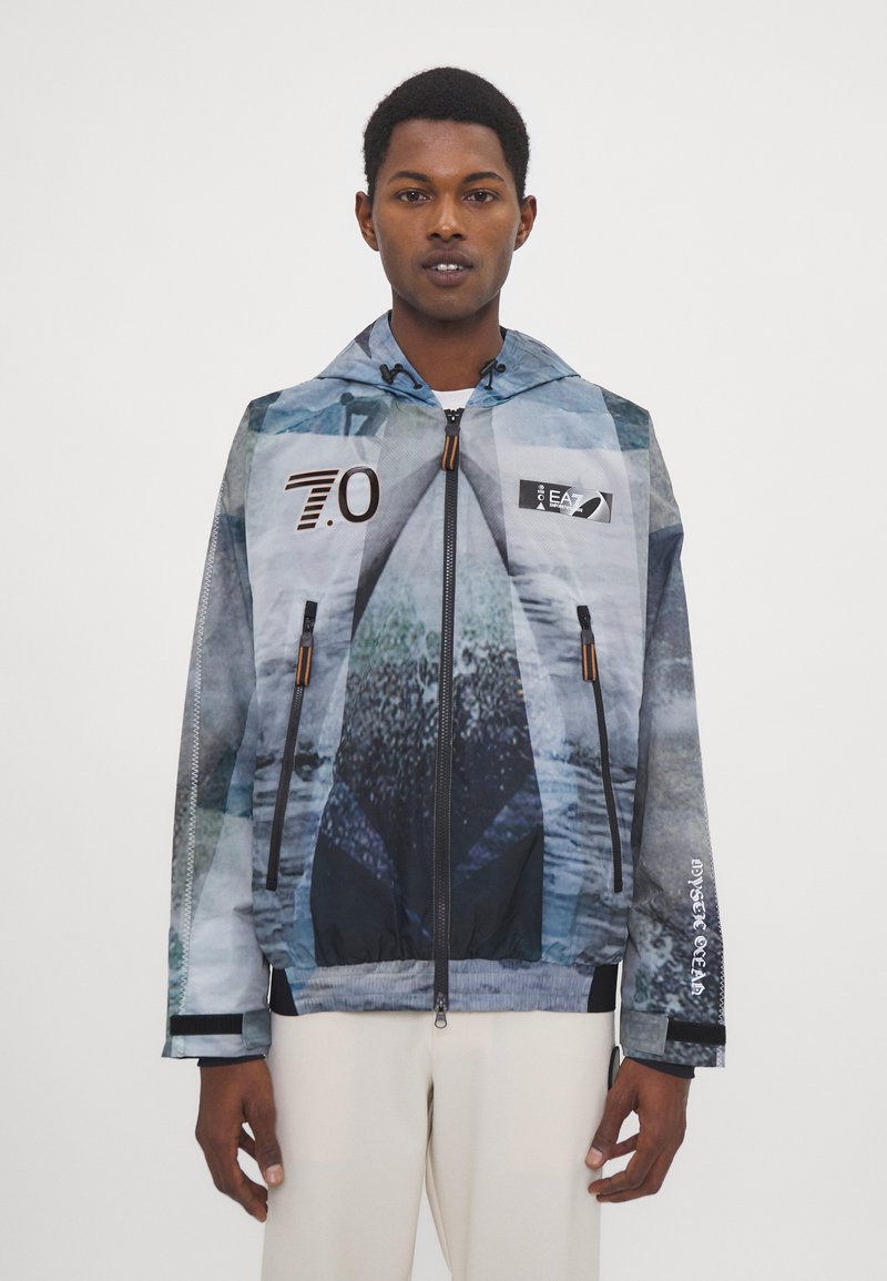 Leichte, mehrfarbige Jacke mit geometrischem Muster, Kapuze, frontalem Reißverschluss und elastischen Bündchen. Verfügt über Logoakzente in kontrastierenden Farben.