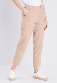 Beige broeken met een ontspannen pasvorm, zijzakken en een gladde textuur, gecombineerd met witte sneakers op een effen achtergrond.