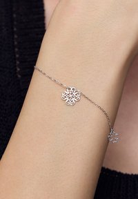 Srebrna bransoletka łańcuszkowa z zawieszką w filigranowym kwiatowym wzorze, z dodatkowym małym akcentem w postaci kwiatu obok zapięcia, spoczywająca na skórze.