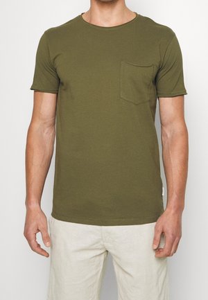 Mann trägt olivgrünes kurzärmeliges T-Shirt mit Brusttasche und hellbeige Hose, steht vor einem einfarbigen Hintergrund.