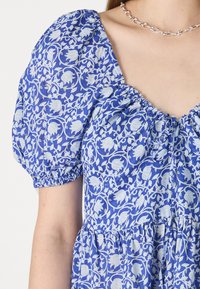 Μπλε φόρεμα με λουλουδένιο σχέδιο, φουσκωτά μανίκια, συγκεντρωμένη λαιμόκοψη και εφαρμοστό κορσέ που μεταβαίνει σε φαρδιά φούστα. Ελαφριά υφή υφάσματος.