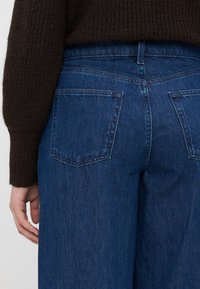 Jeans din denim de culoare albastru închis cu o croială lejeră. Caracteristici includ două buzunare la spate și un talie înaltă. Țesătura texturată este vizibilă.