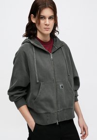 Sudadera gris con cremallera, bolsillo frontal, capucha con cordón y un parche de logo. Fabricada en tejido suave, presenta un ajuste relajado y puños acanalados.