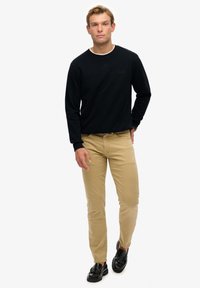 Sort sweatshirt med rund hals, khaki slim-fit bukser og sorte loafers med sløjfedetalje. Modellen står foran en ensfarvet hvid baggrund.