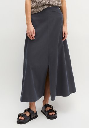 Persona che indossa una gonna midi grigio scuro con uno spacco frontale e sandali neri con fibbie dorate, in piedi contro uno sfondo semplice.