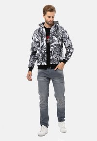 Schwarz-weiße gemusterte Zip-Hoodie mit Kordelzugkapuze, gerippten Bündchen und Saum. Getragen über einem schwarzen Grafik-T-Shirt und grauen Jeans.