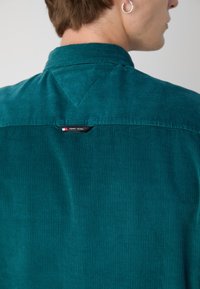 Camisa de pana azul verdoso con cuello de camisa, cuenta con un yugo triangular y una etiqueta visible de "TOMMY JEANS" en la parte trasera.