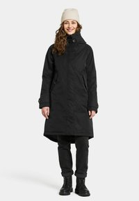 Zwarte lange jas met een hoge kraag, voorrits en zijzakken. Gecombineerd met zwarte jeans en stevige zwarte veterschoenen.