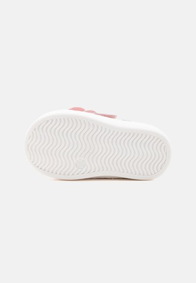 Semelle en caoutchouc blanc d'une basket enfant avec un motif ondulé et un logo circulaire près du talon, vue par le dessous.